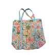 A canvas tote bag featuring a colorful tropical print with palm trees, hibiscus flowers, and sun motifs in pink, orange, green, and blue, complete with braided blue and white shoulder straps.