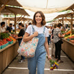 A woman walking through a bustling outdoor farmers market wearing light blue denim overalls and a white t-shirt, carrying a large floral print tote bag and holding a small bunch of colorful wildflowers.