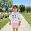 A young child standing on a paved path in a sunny park, wearing a white long-sleeve top with a colorful unicorn and rainbow graphic, paired with light pink shorts.