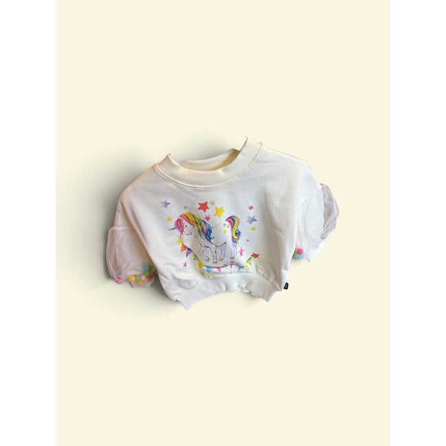 A white cropped sweatshirt featuring a colorful graphic of a unicorn with a rainbow mane, surrounded by stars, displayed against a solid cream-colored background. Pom poms in the tulle sleeves.