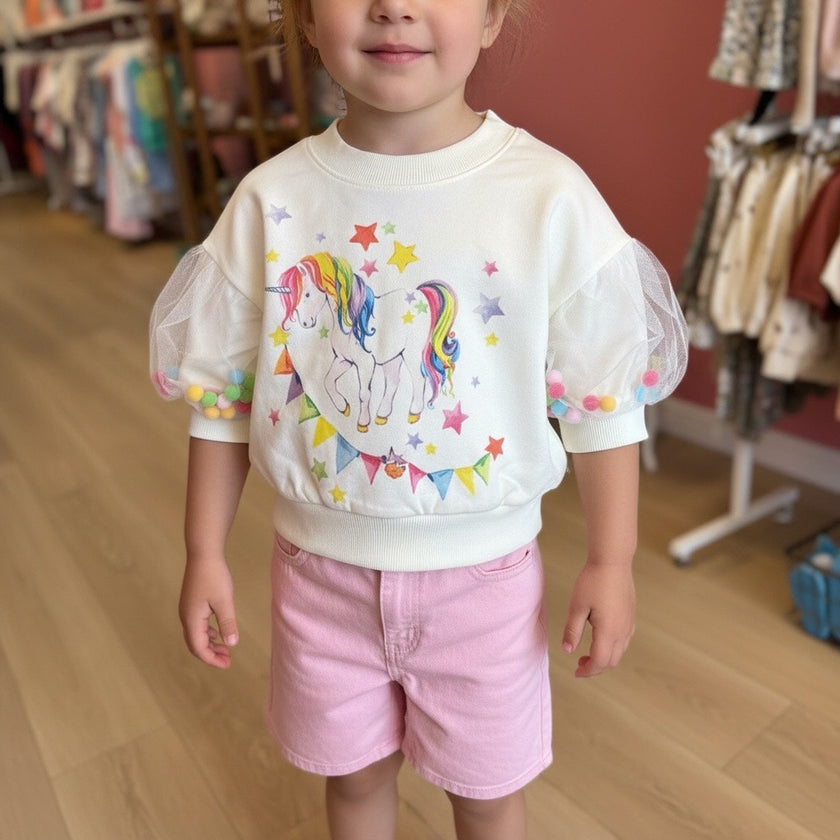 A toddler wearing a cream-colored sweatshirt with a colorful rainbow unicorn graphic, sheer puff sleeves with pom pom inside, and pink shorts, standing in a clothing store.